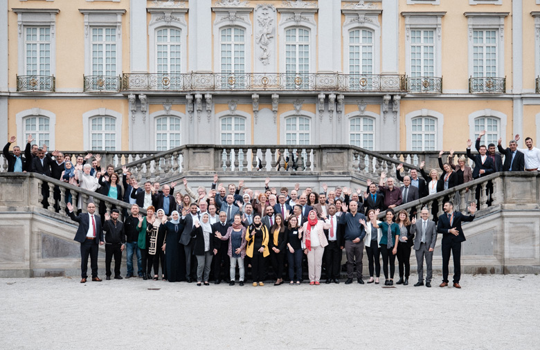 The photo shows the participants of the 4th German-Palestinian Partnership Conference on and in front of the steps of a staircase that leads to the right and left to a raised level in front of the Brühl Castle. In the background you can see the window front and the yellow baroque façade of Augustusburg Palace.